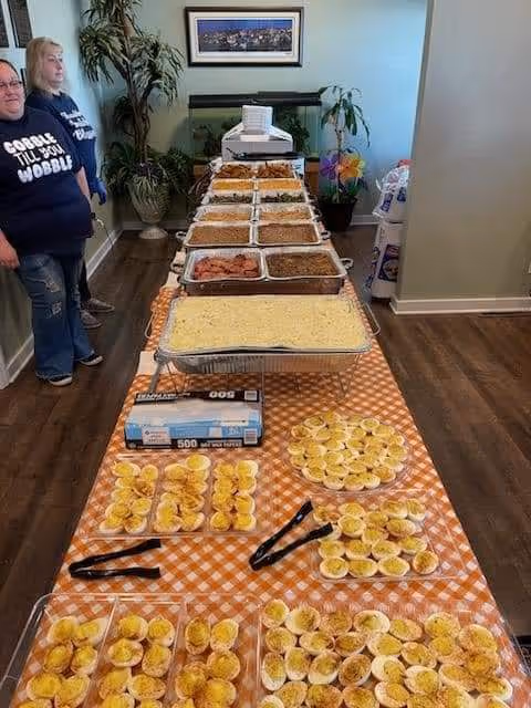 Long buffet line with trays of food in a dining area; two people stand nearby.
