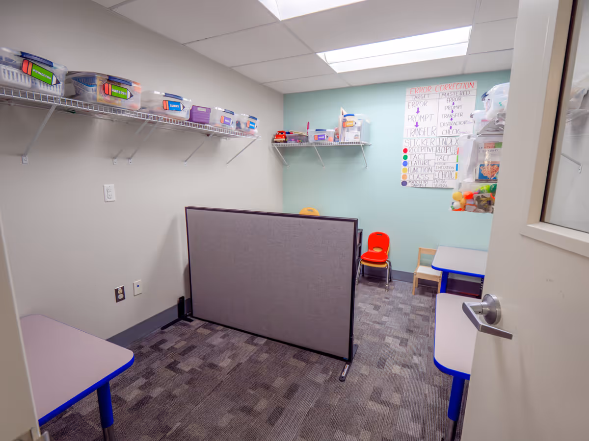 A small, organized activity room with labeled bins, a whiteboard, and low tables.
