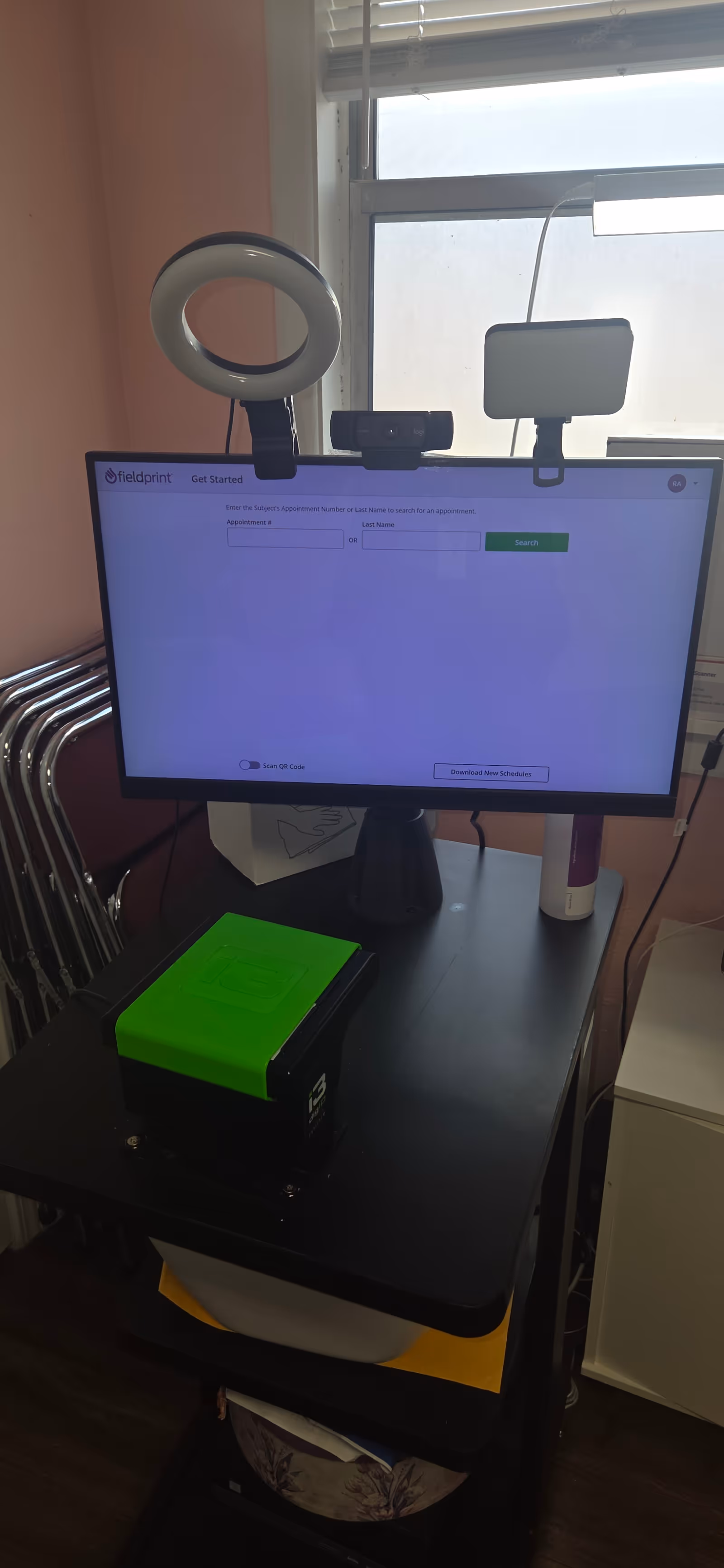 A computer workstation on a mobile cart with monitor, webcam, ring light, and green device.