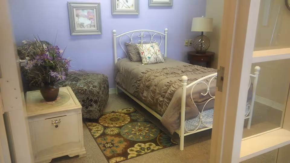 A cozy bedroom in a senior facility with a white metal bed, nightstand, and rug.