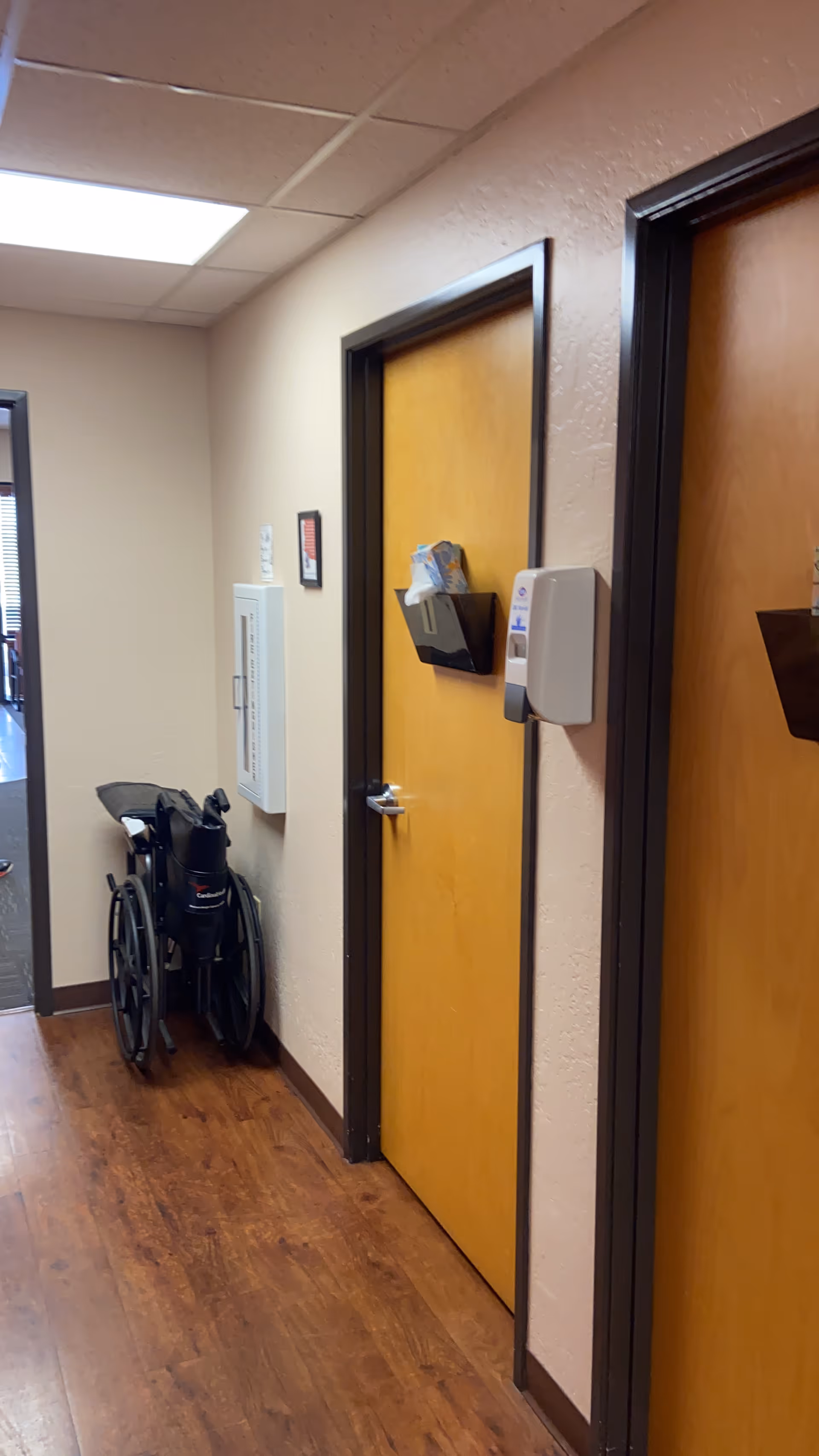 Wheelchair sits in a hallway beside wooden doors and wall-mounted supplies.
