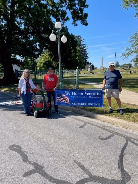 Three people stand outdoors beside a Geisinger Hospice banner honoring veterans.