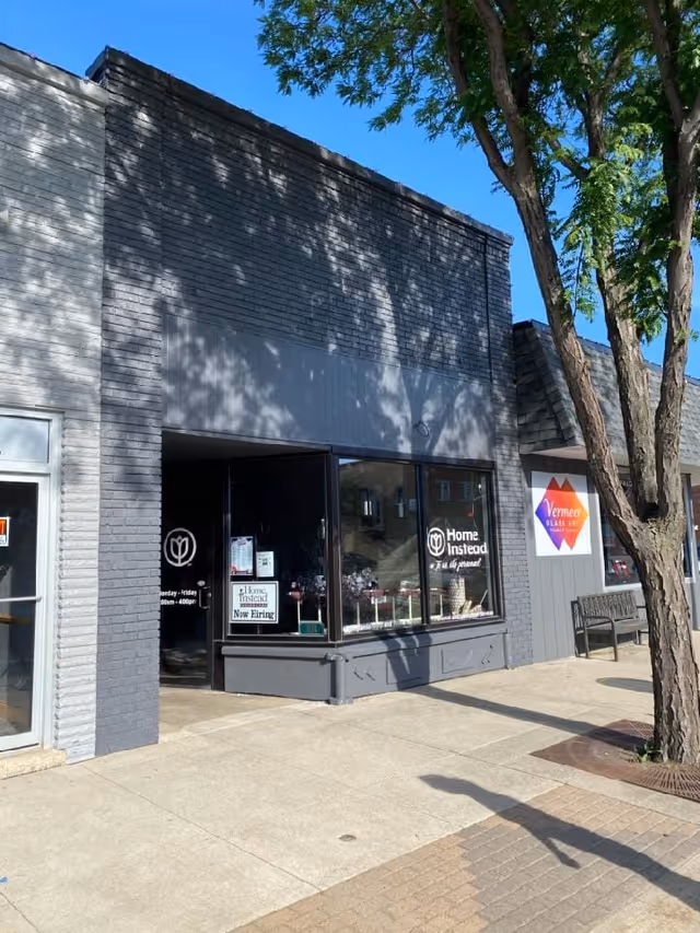Front exterior of a Home Instead office storefront with display windows and a bench nearby