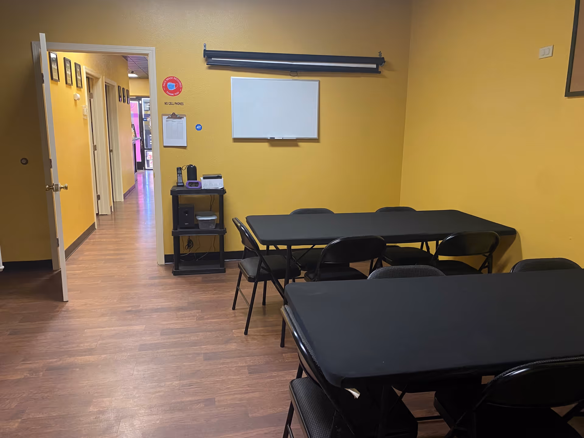 Yellow-walled interior room with black tables and chairs, whiteboard, and hallway.