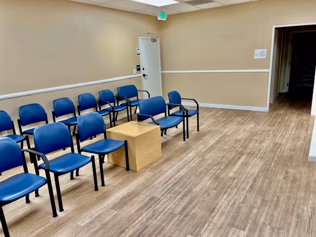 Beige-walled interior waiting area with blue chairs arranged in rows and a small wooden table.
