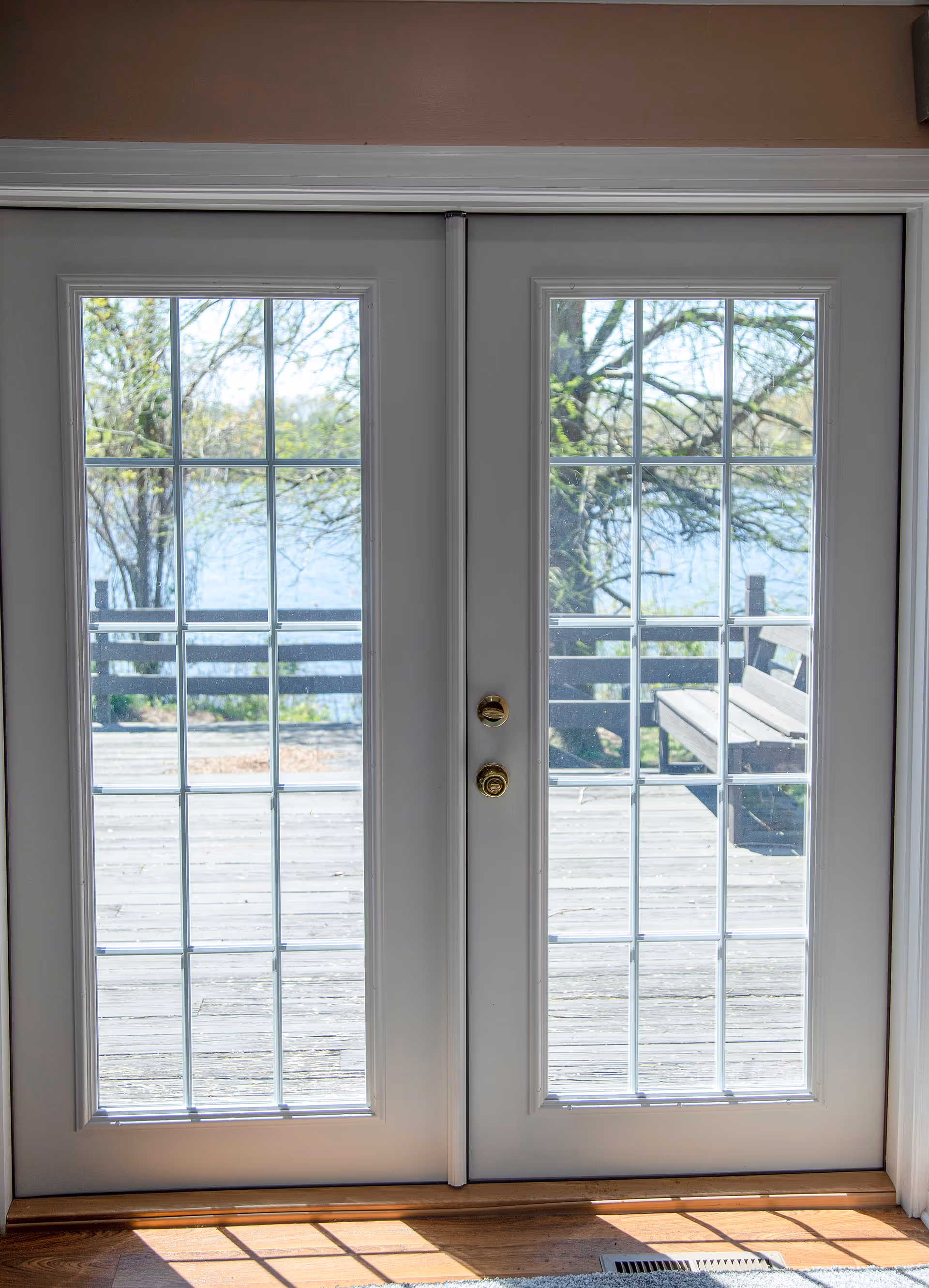 Double glass doors open to a wooden deck with trees and a bench outside.