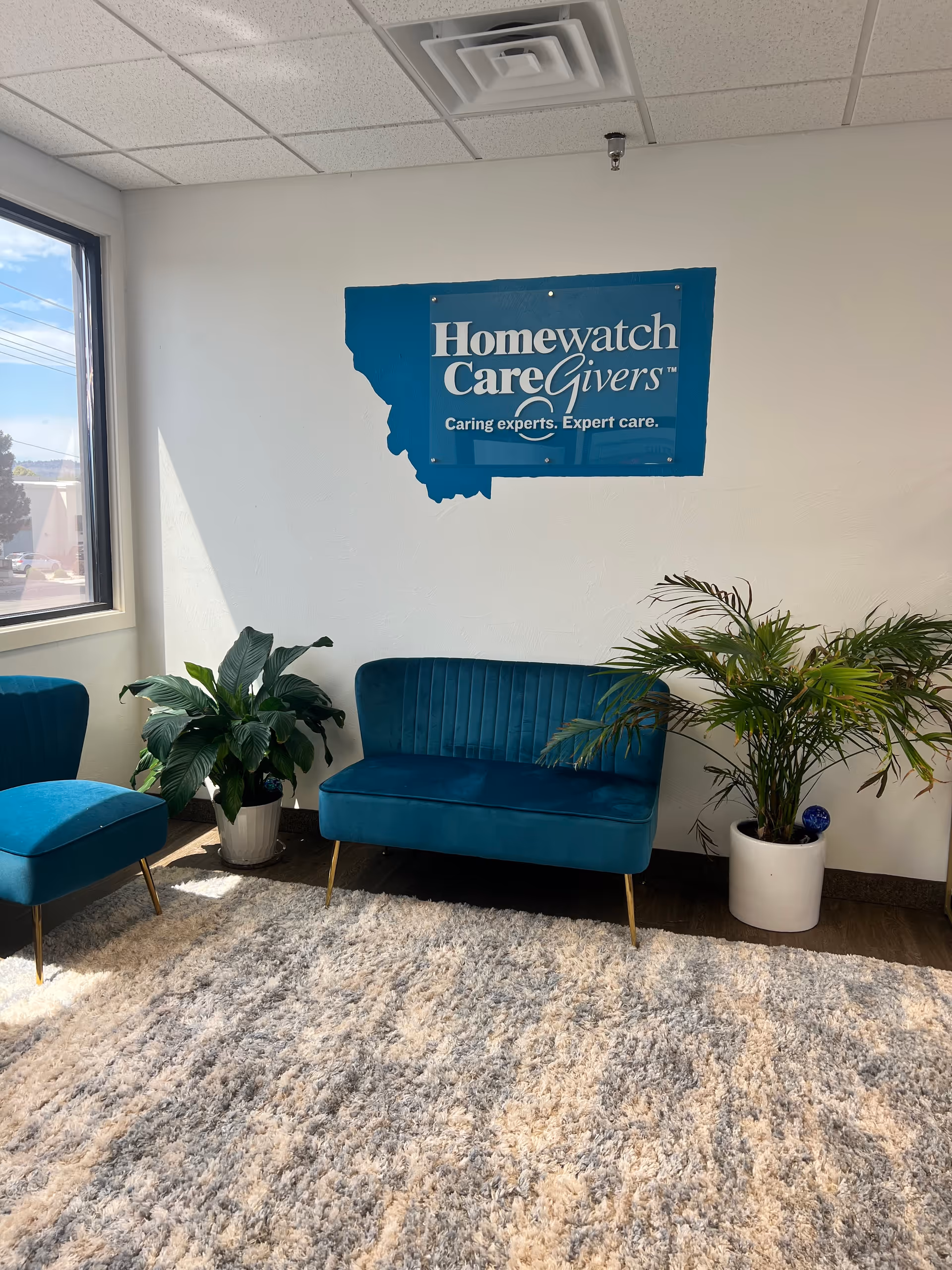 Lobby seating area with teal furniture, plants, and a Homewatch CareGivers banner.
