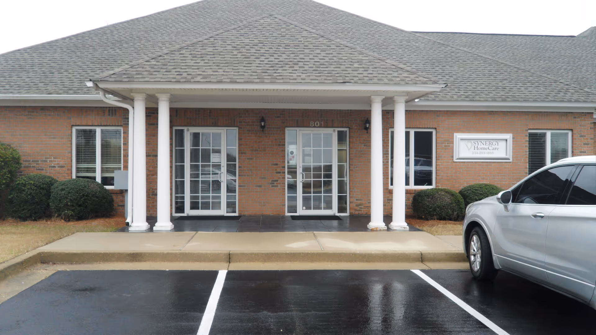 Brick senior living entrance with white columns, glass doors, and a parked silver car in front.