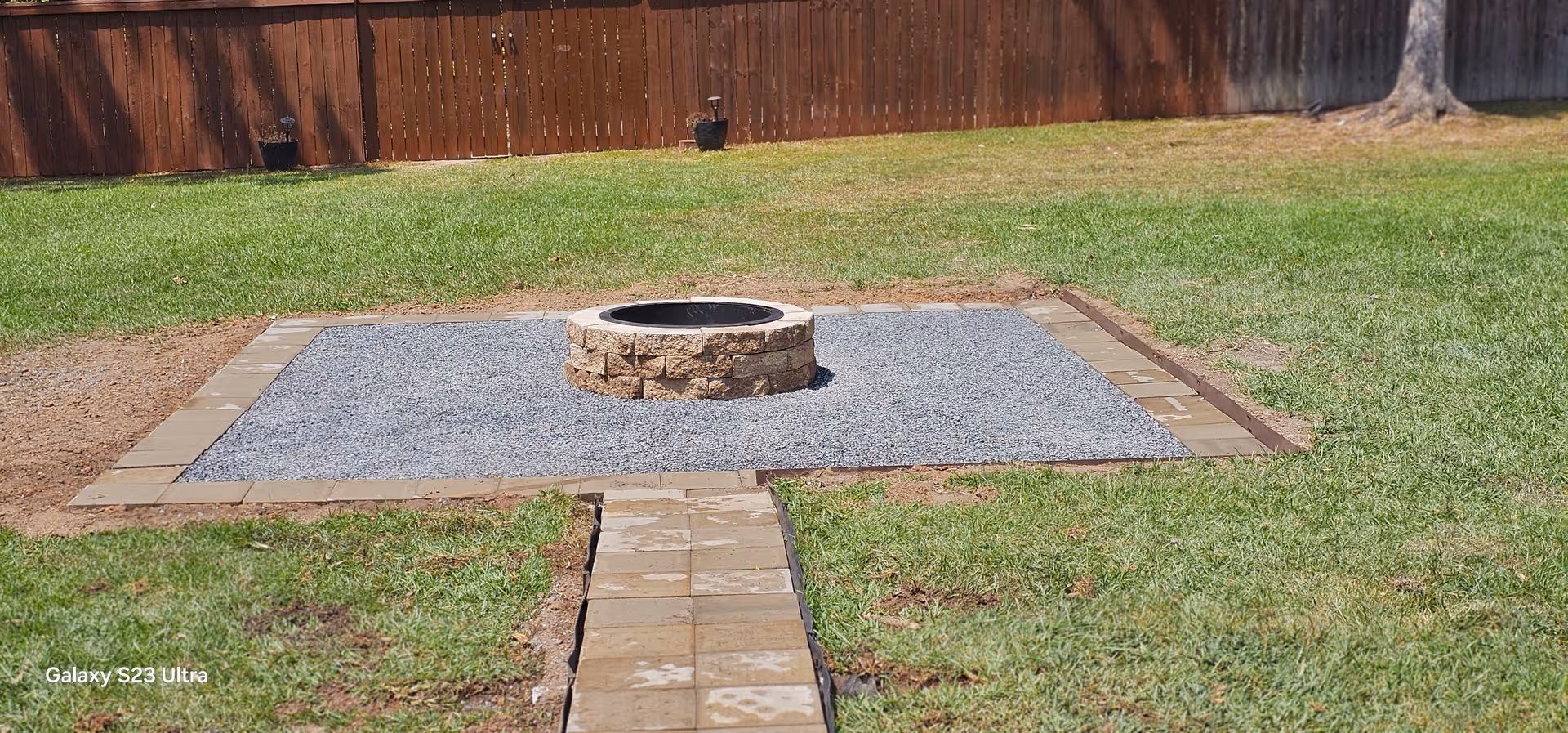 Backyard with circular stone fire pit on gravel pad and brick path.