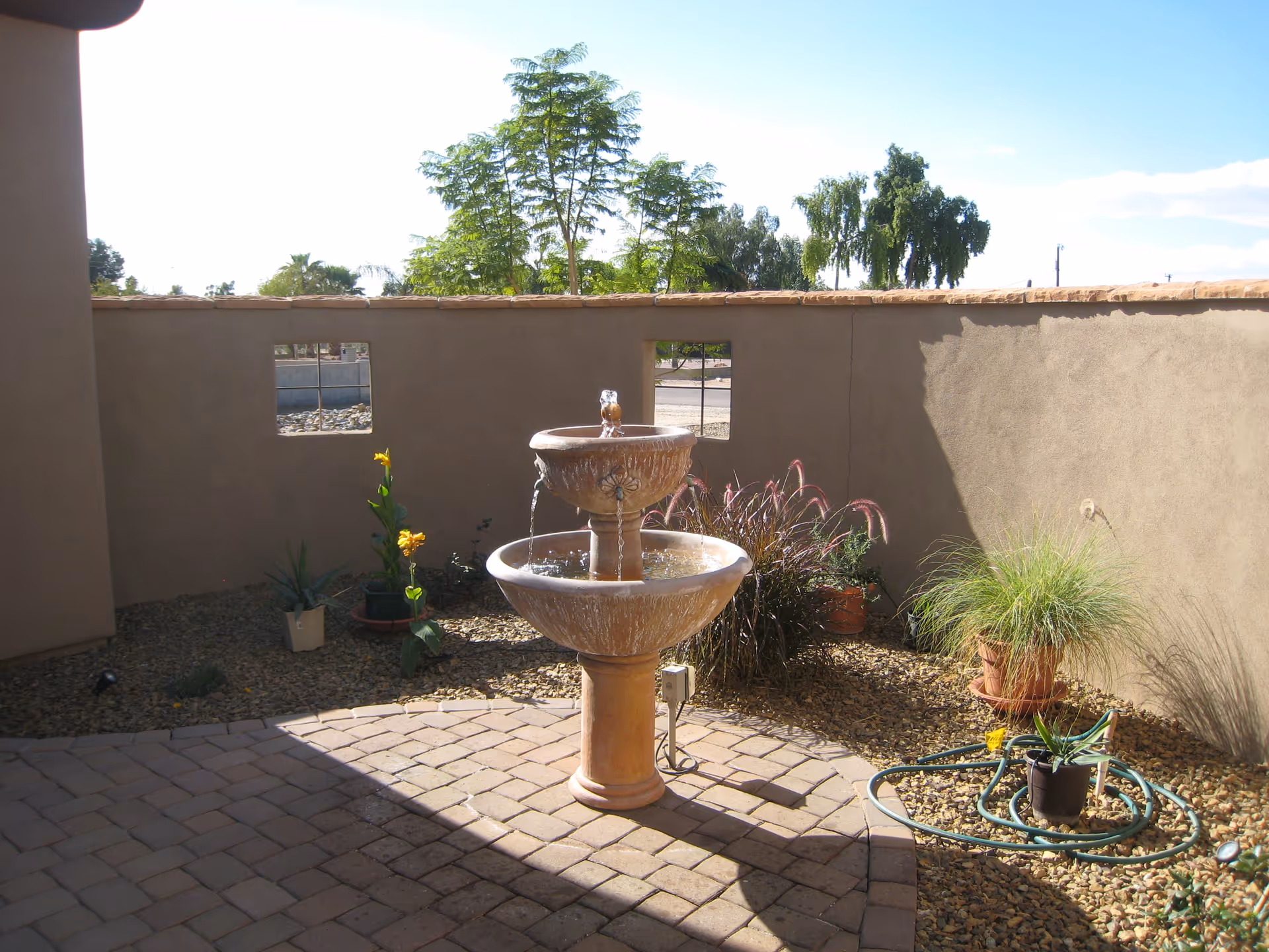 Sunlit courtyard with tiered fountain, potted plants, and a gravel garden area.