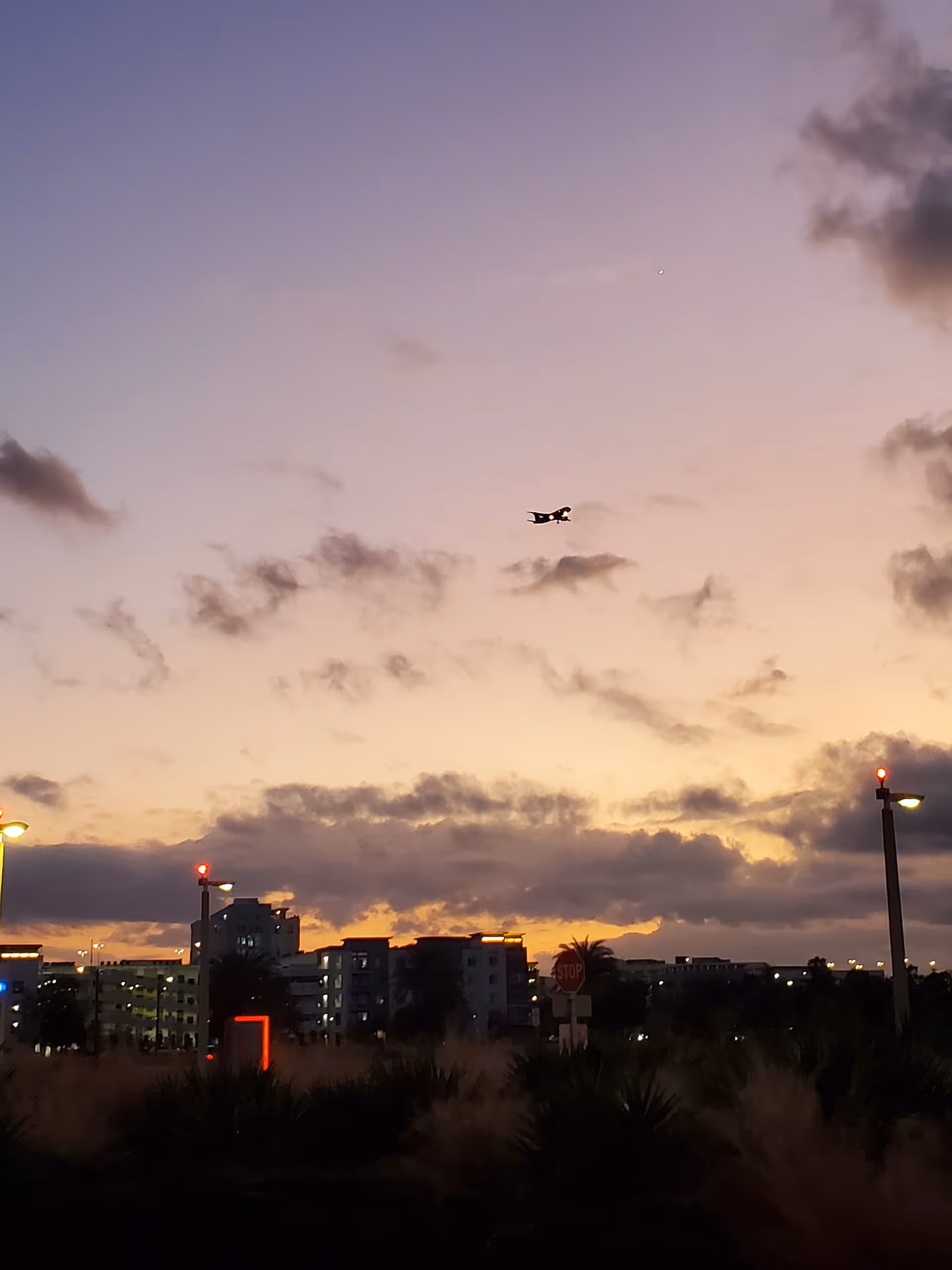 Sunset sky over a campus with buildings, streetlights, and a distant airplane.