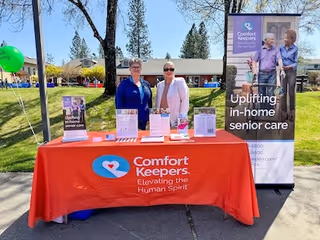 Outdoor booth featuring a red Comfort Keepers tablecloth, banner, and two staff members.