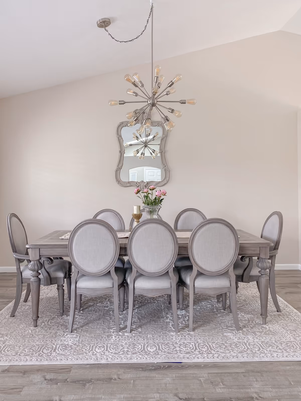 A grey wooden dining table with eight upholstered chairs, chandelier, mirror, and flowers.