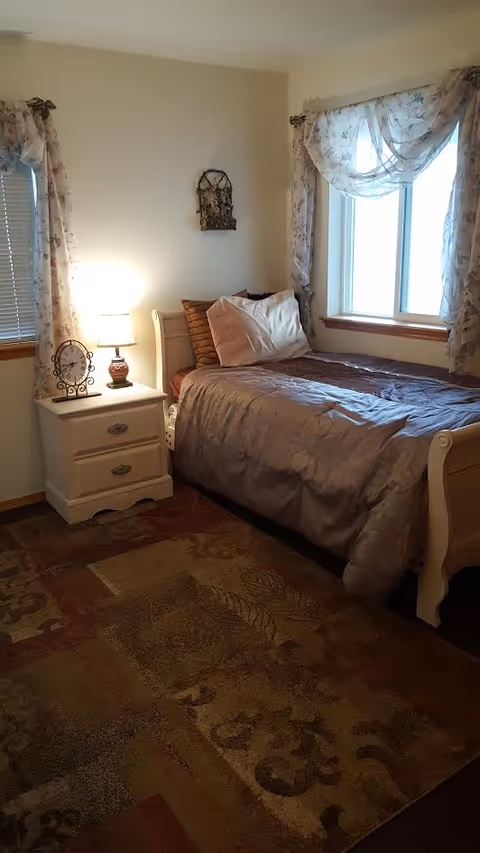 Cozy bedroom with a bed, nightstand, lamp, and floral curtains by windows.