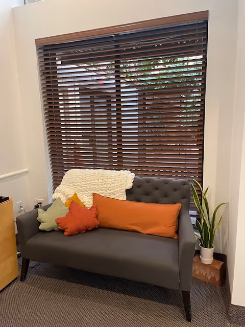 A grey sofa with colorful cushions and a white throw by a window with blinds.