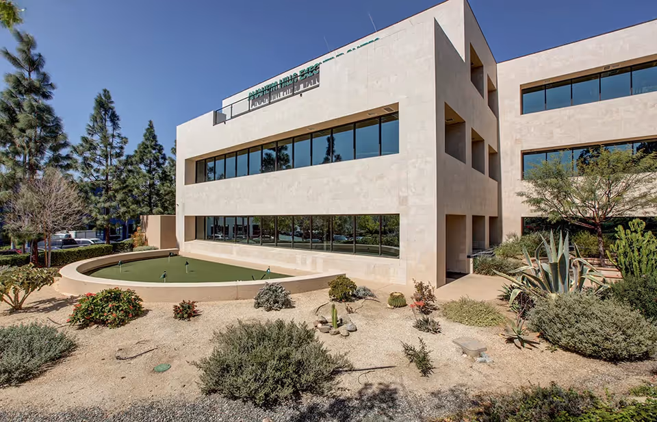 Modern beige building exterior with large windows and a circular fountain in front.