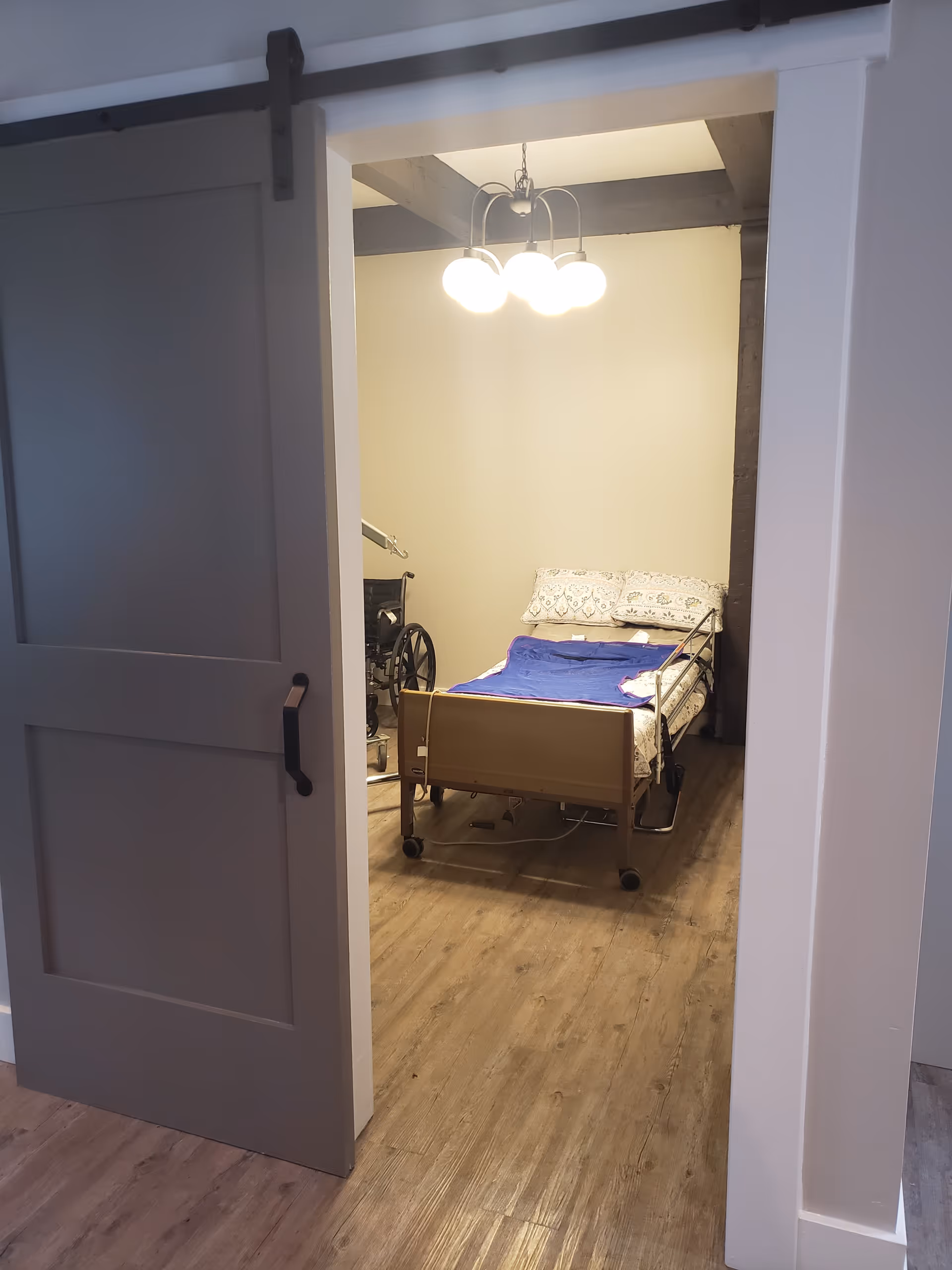 View into a senior living bedroom with a hospital bed and wheelchair near the wall.