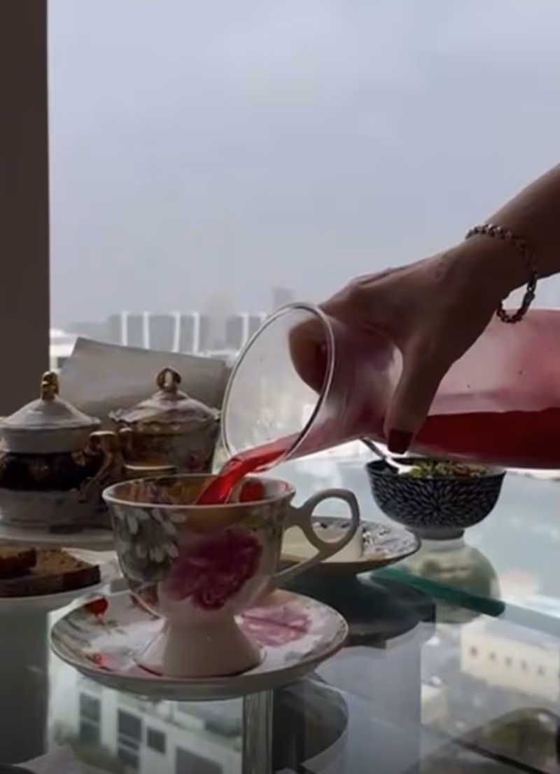 A hand pours pink juice into a floral teacup on a glass dining table.