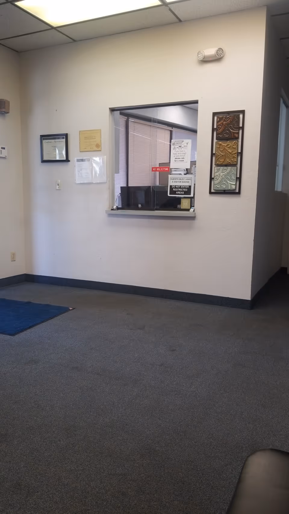 Interior reception area with a windowed counter, wall certificates, and carpeted floor.