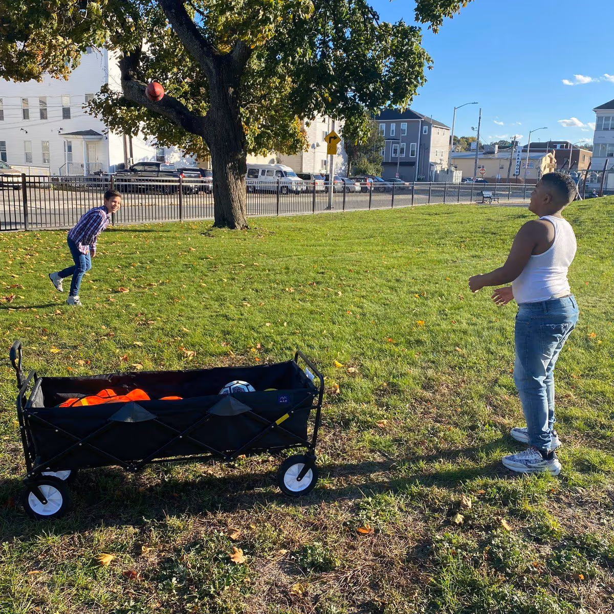 Two children play with a wagon and ball in a sunny grassy park.