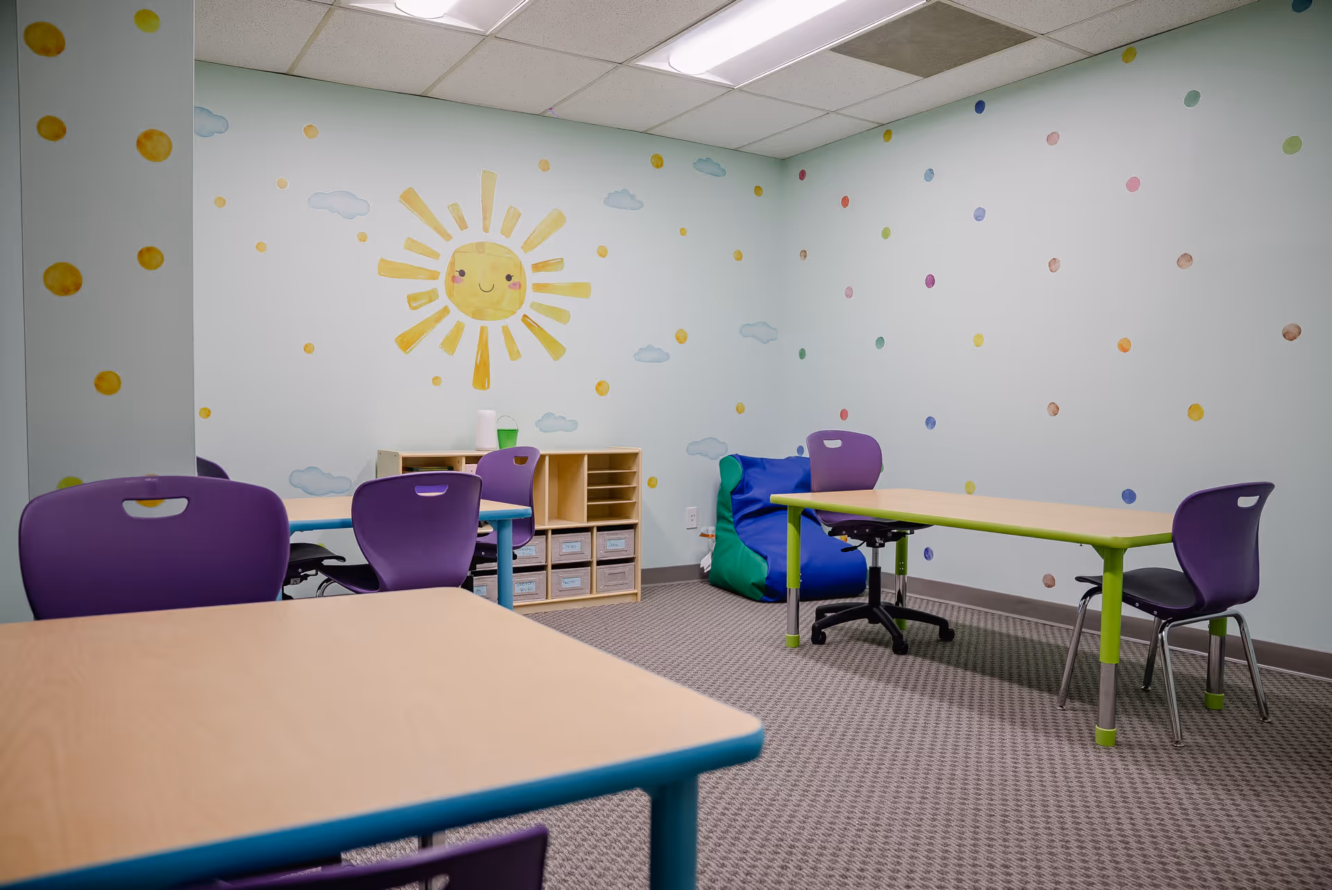 Bright therapy room with pastel walls, sun mural, child-sized tables, and purple chairs.