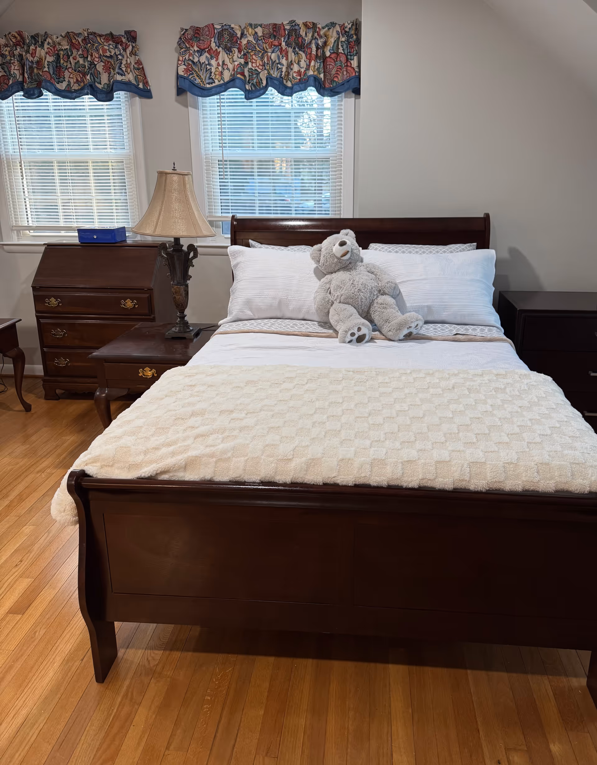 A neatly made bedroom with dark wood furniture, windows, and a teddy bear on the bed.