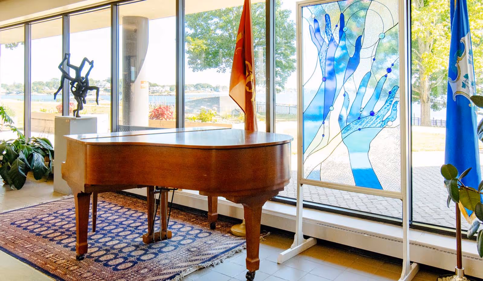 A wooden grand piano sits on a rug beside bright windows and artwork.