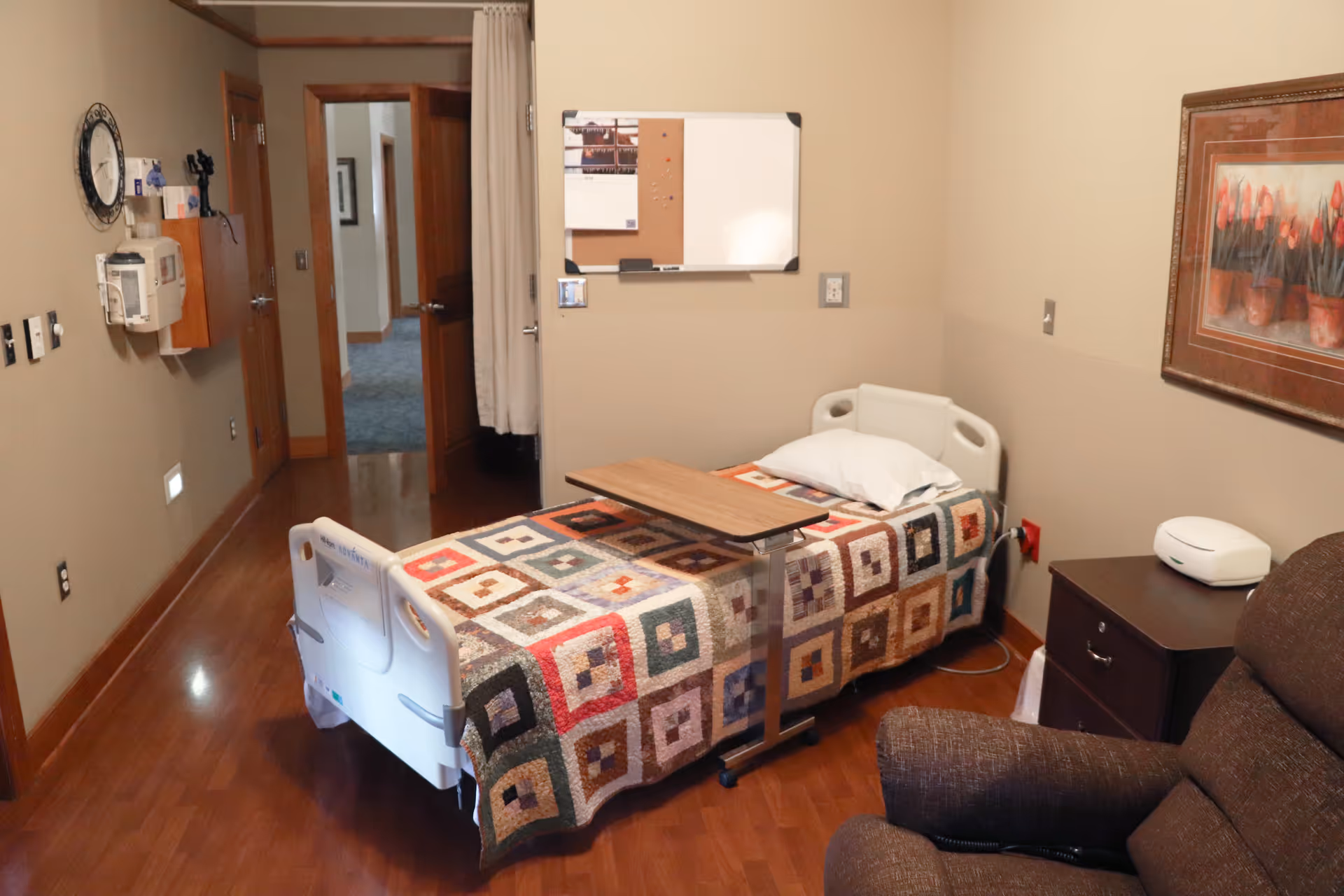 A patient room with single bed, patterned quilt, wood floor, and a brown recliner.