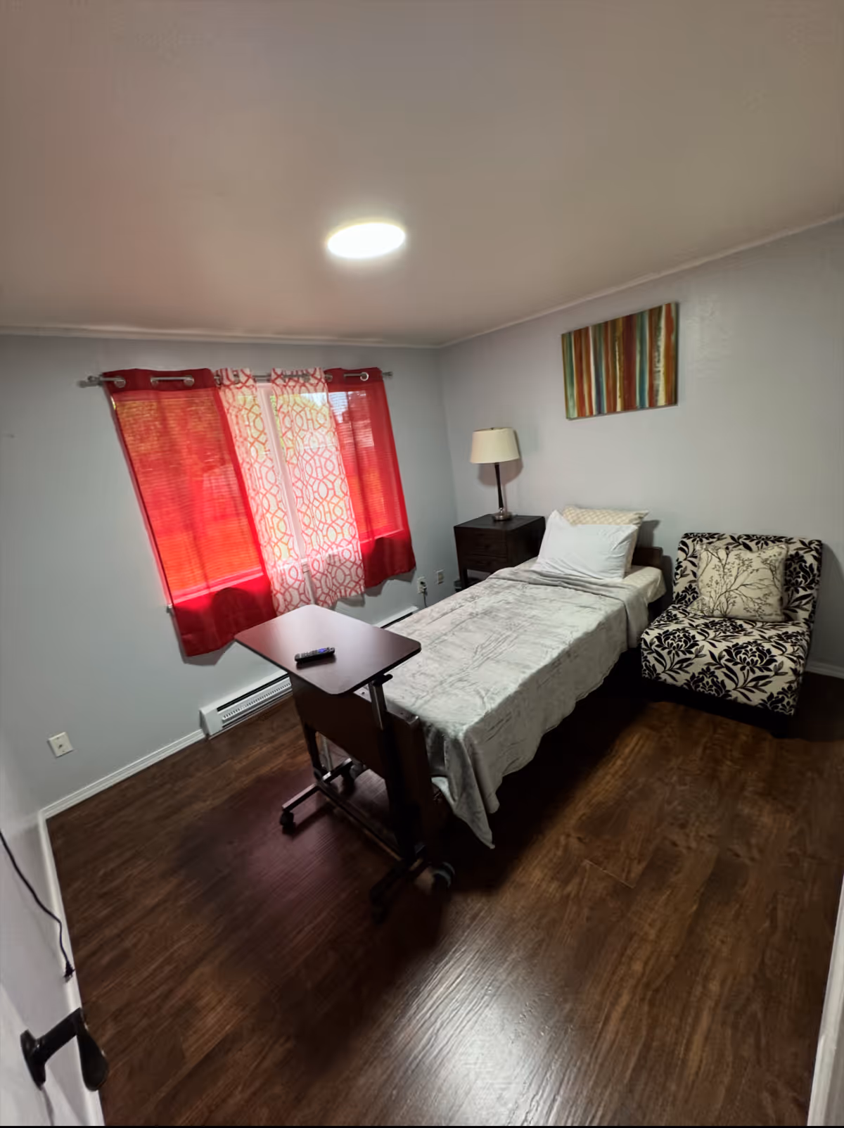 Small bedroom with a single bed, chair, nightstand, lamp, and red curtains.