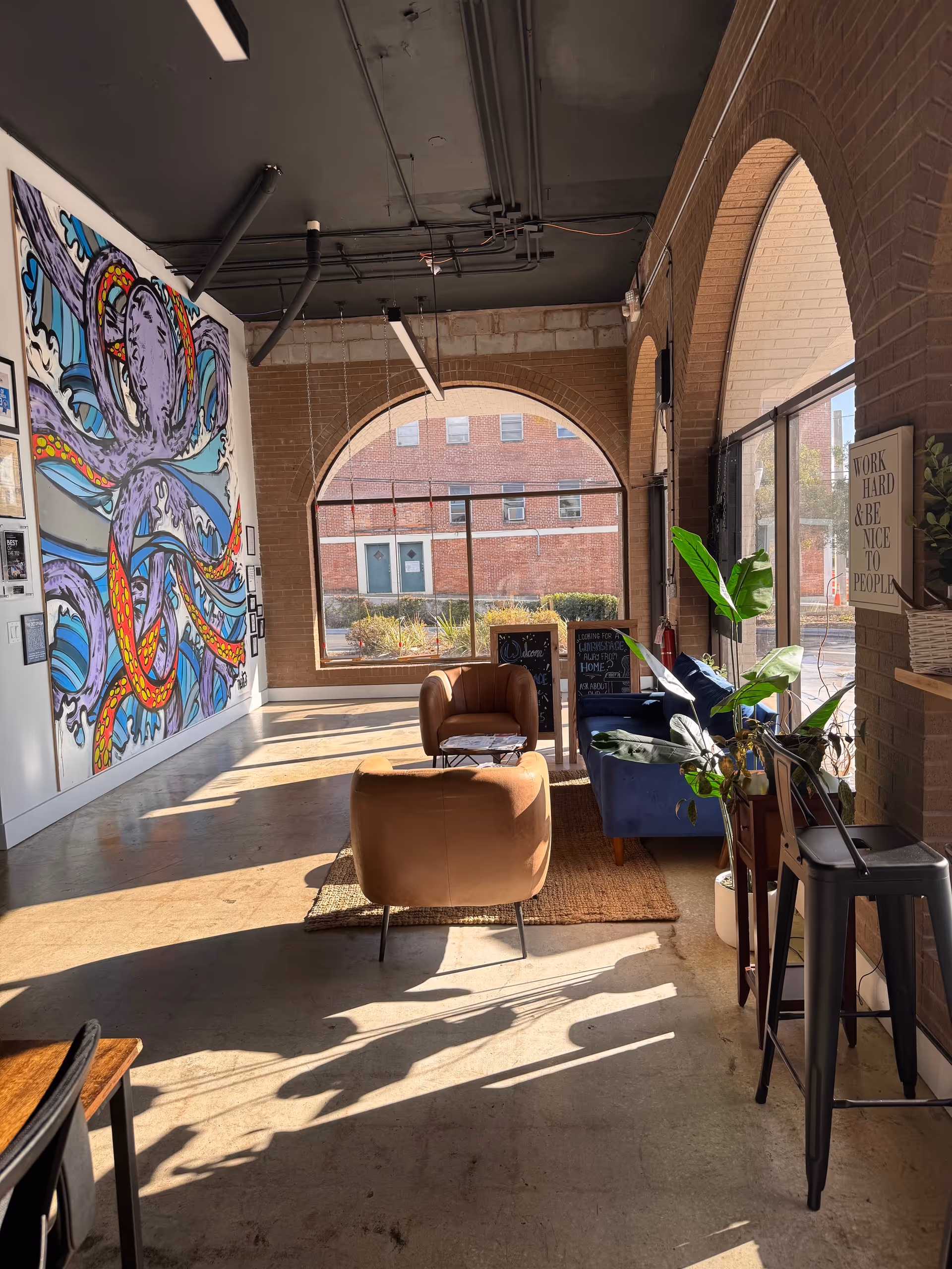 Sunlit industrial-style lounge with arched windows, colorful mural, and cozy seating.