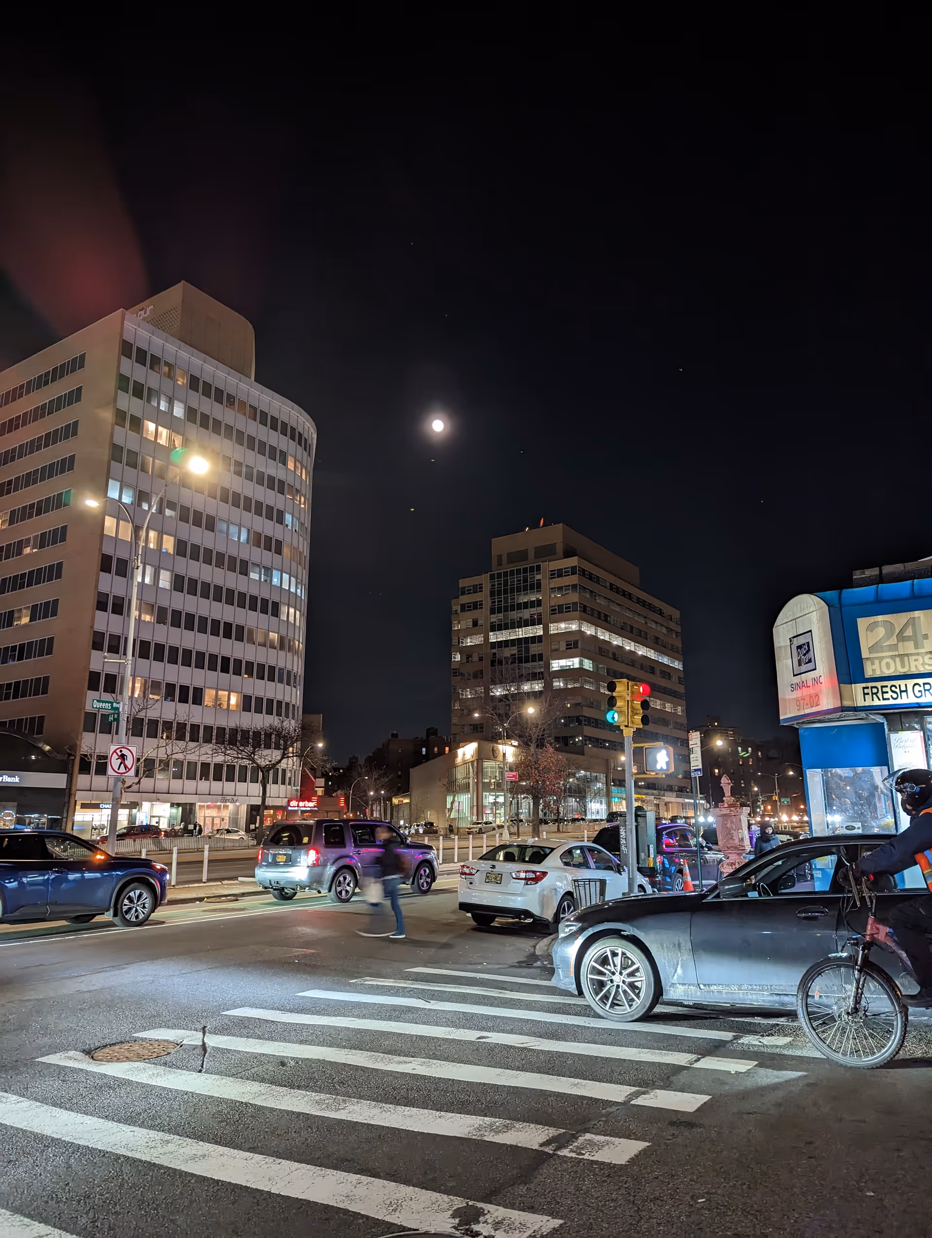 Nighttime city street with crosswalk, vehicles, a cyclist, and tall office buildings.