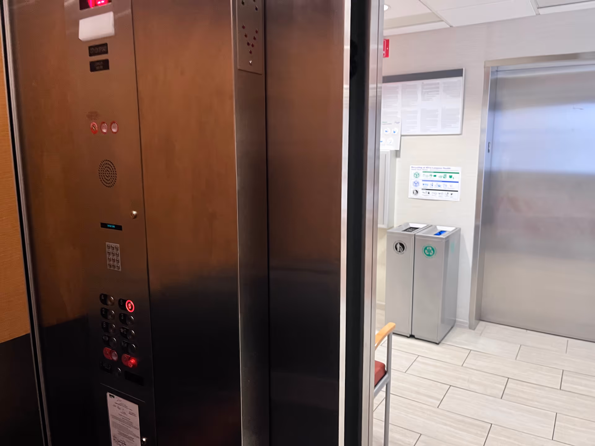 Stainless steel elevator doors in a clinical lobby with a control panel and waste bins.