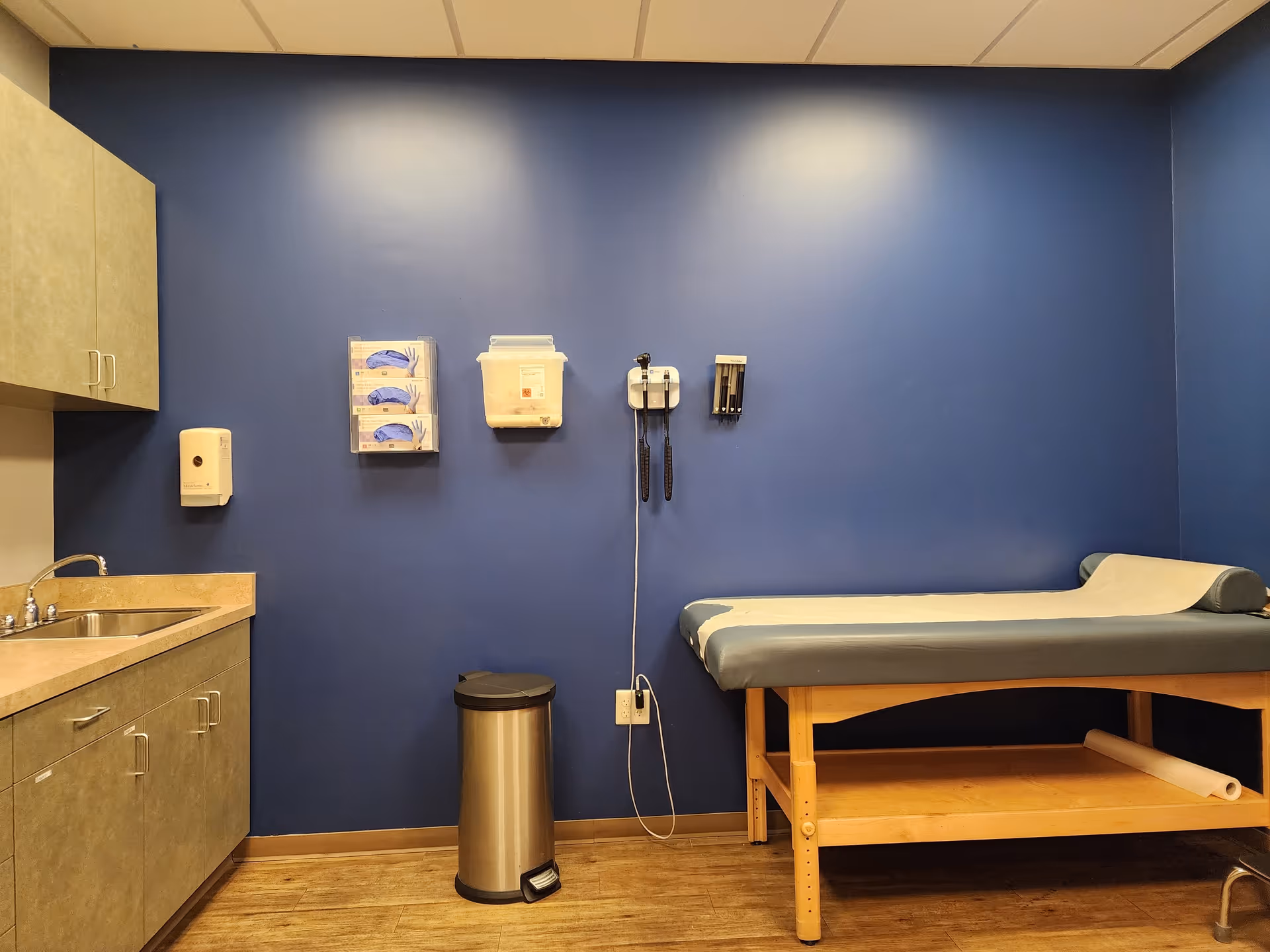 Blue-walled medical examination room with sink, cabinets, and an exam table.
