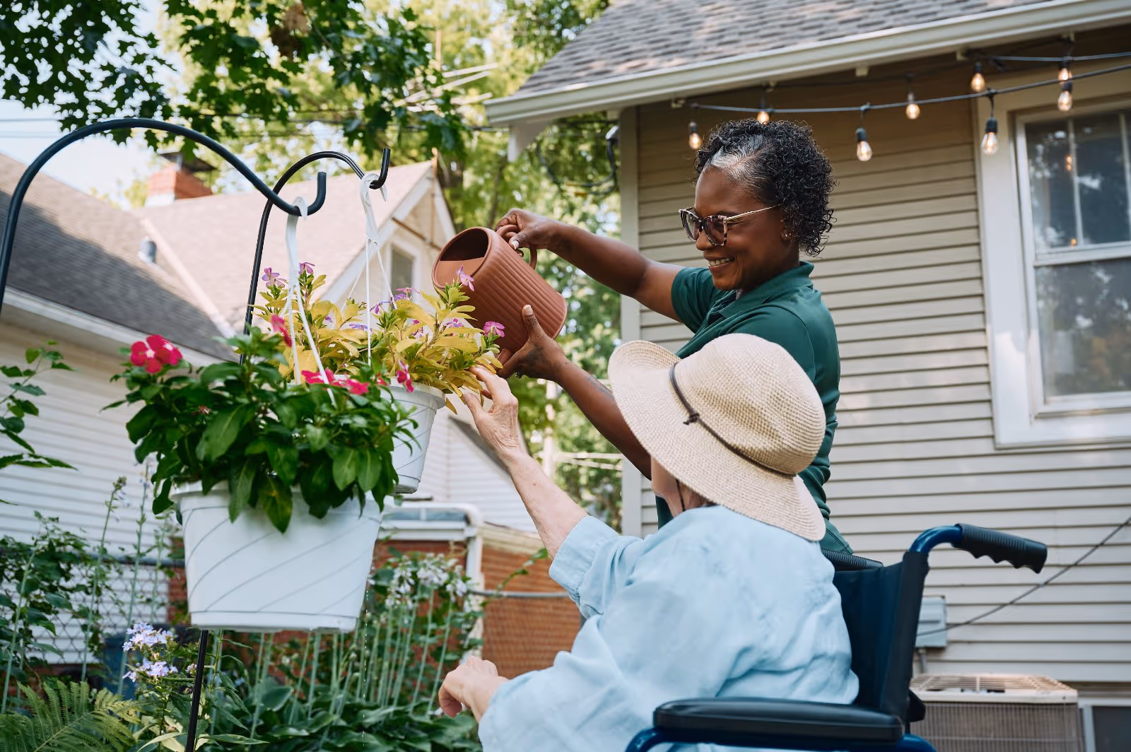 Two caregivers assist an elderly resident watering hanging flower baskets in an outdoor garden.