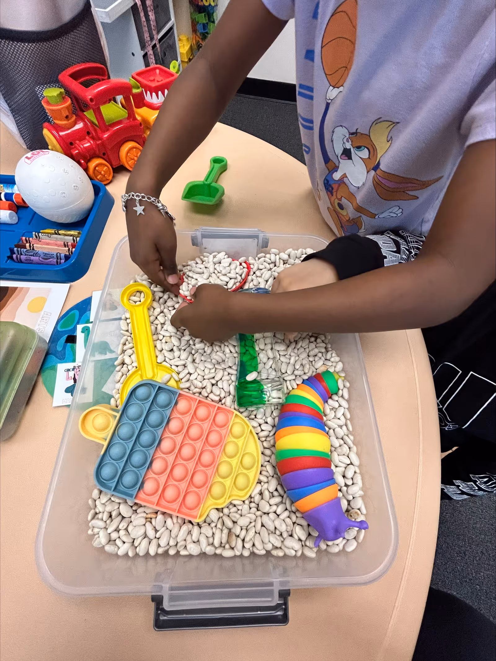 Hands of a person play with a sensory bin filled with white pebbles and colorful toys.
