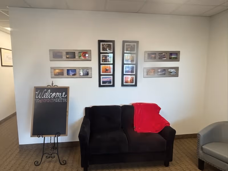 Lobby seating area with black sofa, red blanket, framed wall photos, and a welcome board.