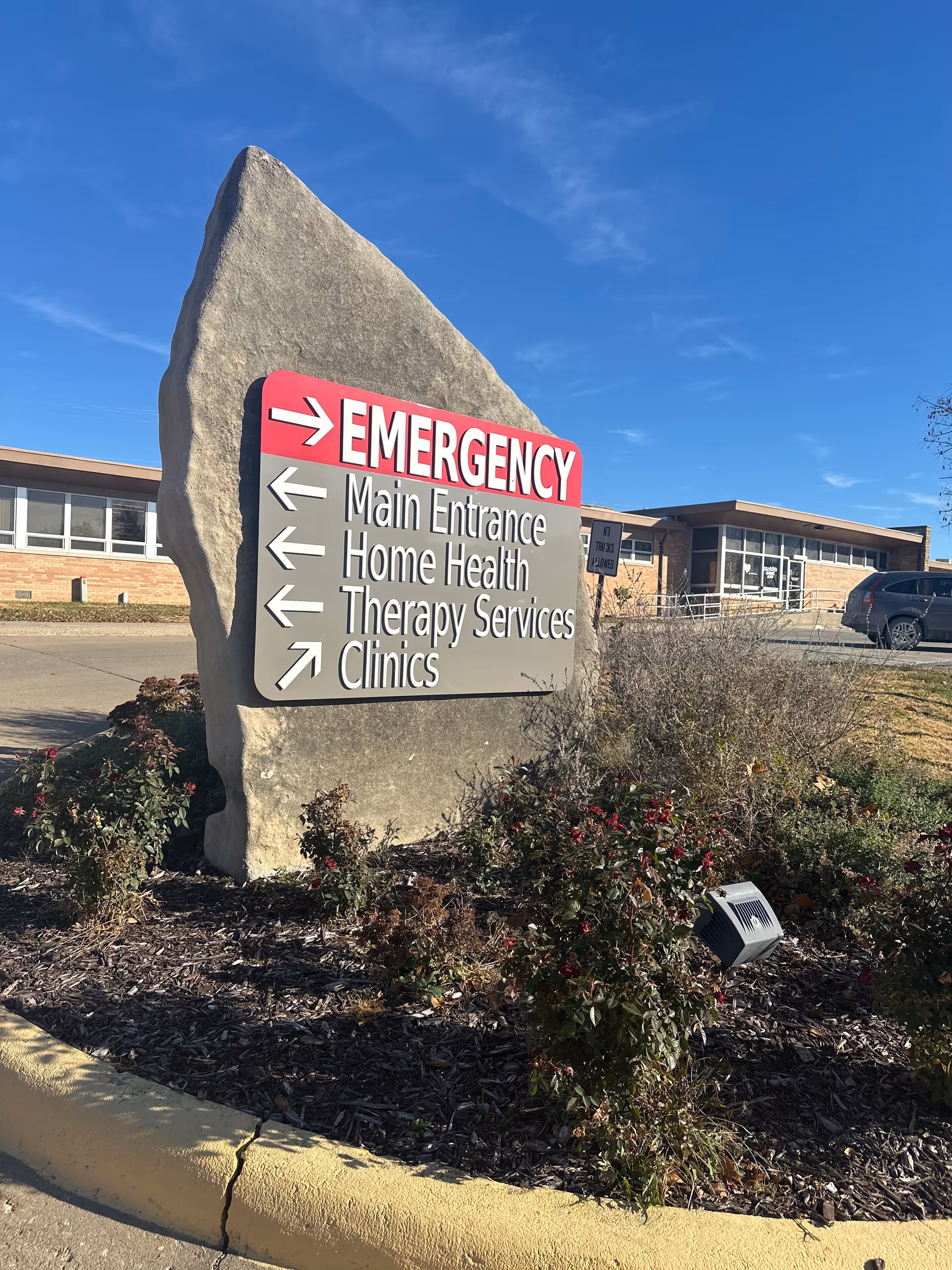 A tall rock sign with an Emergency sign and arrows near a hospital entrance.