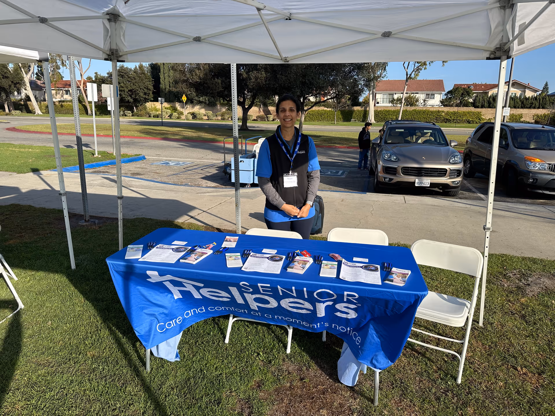 A caregiver stands under a white canopy beside a blue Senior Helpers table outside.