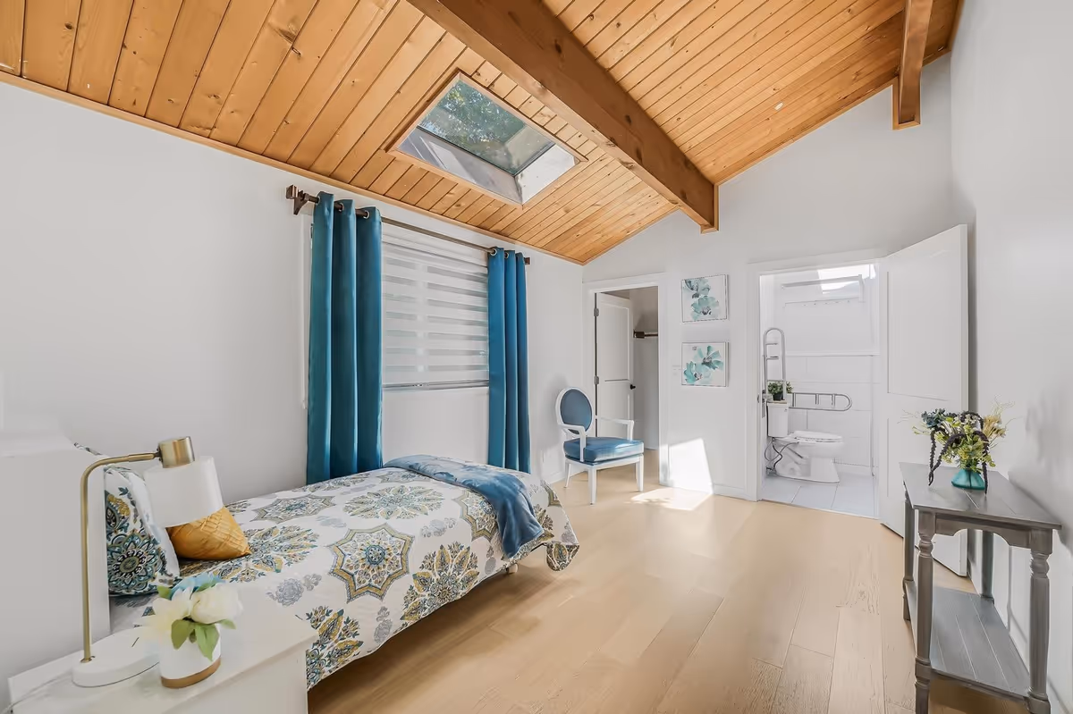 Bright bedroom with patterned bed, teal curtains, skylight, and adjacent bathroom.