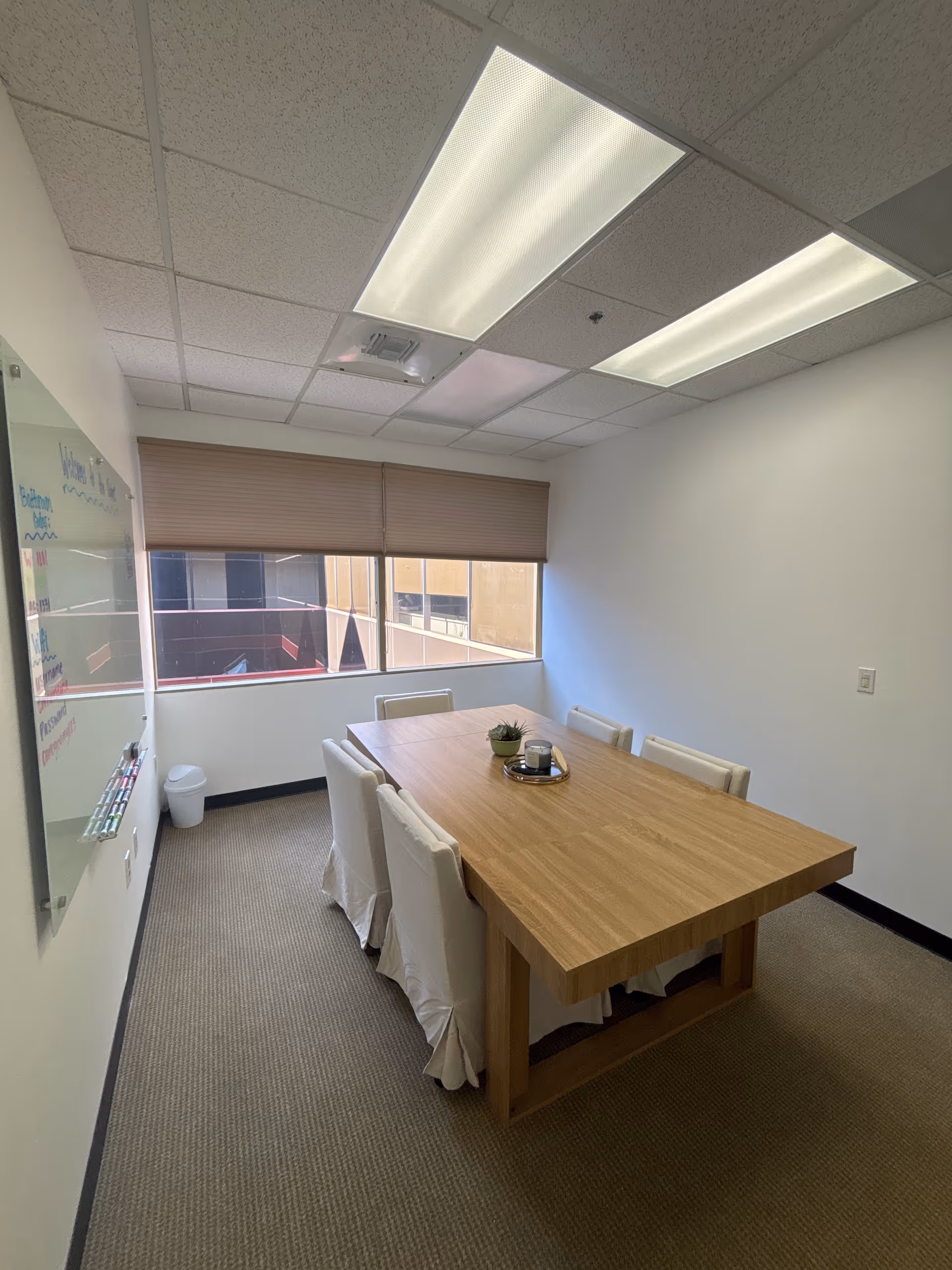 A modern meeting room with a wooden table, white chairs, whiteboard, and window blinds.