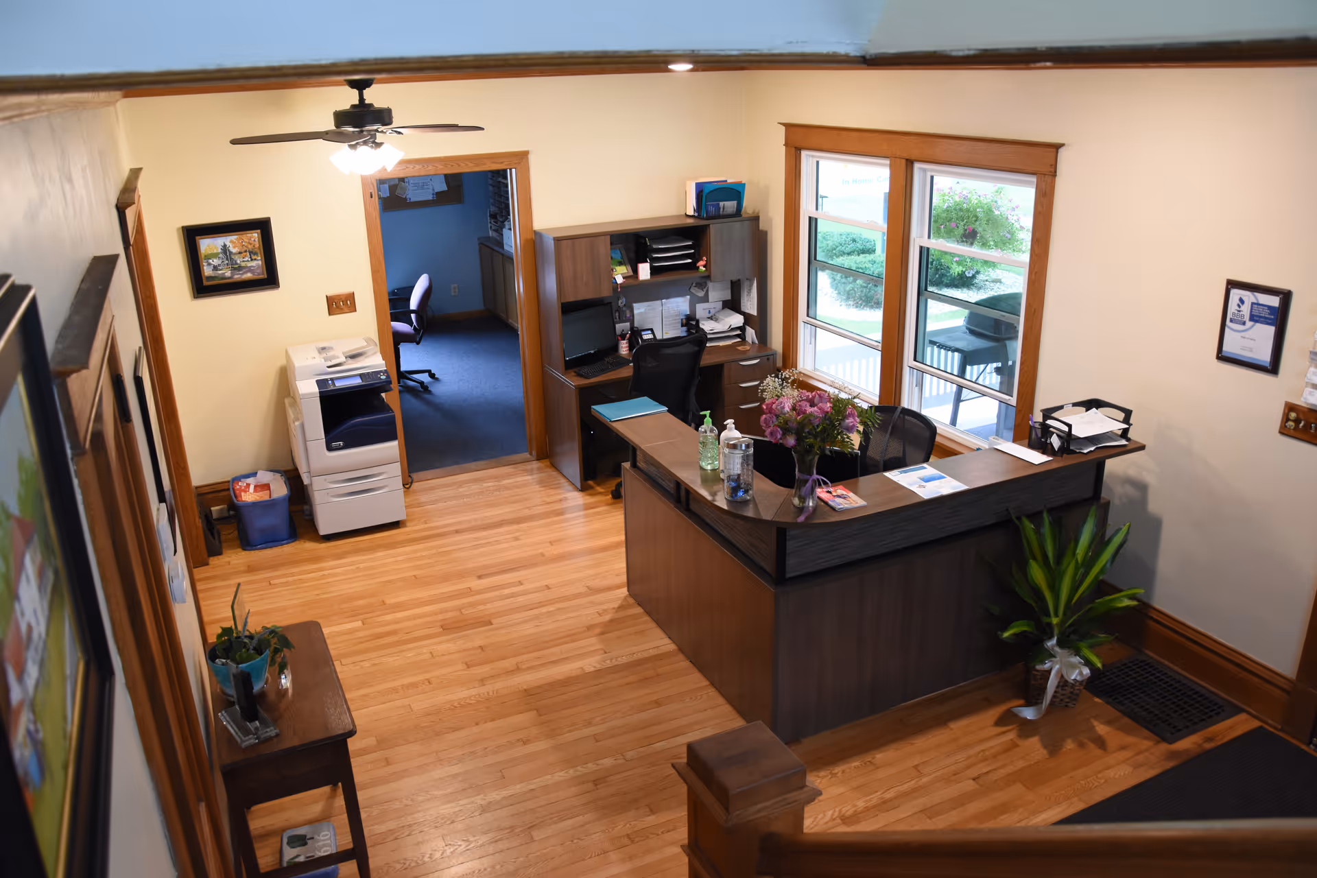 Reception desk in a senior living lobby with wood floors, windows, plants, and chairs.