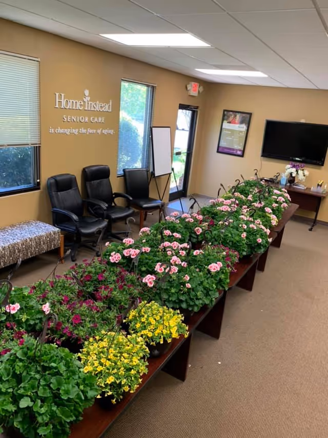 Inside a Home Instead Senior Care lobby, row of black chairs and flowering plants line hallway.