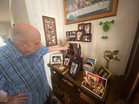 Elderly man in plaid shirt points at framed photos on a wall display.