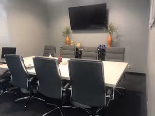 A conference room with a long table, surrounding chairs, a wall TV, and decorative plants.