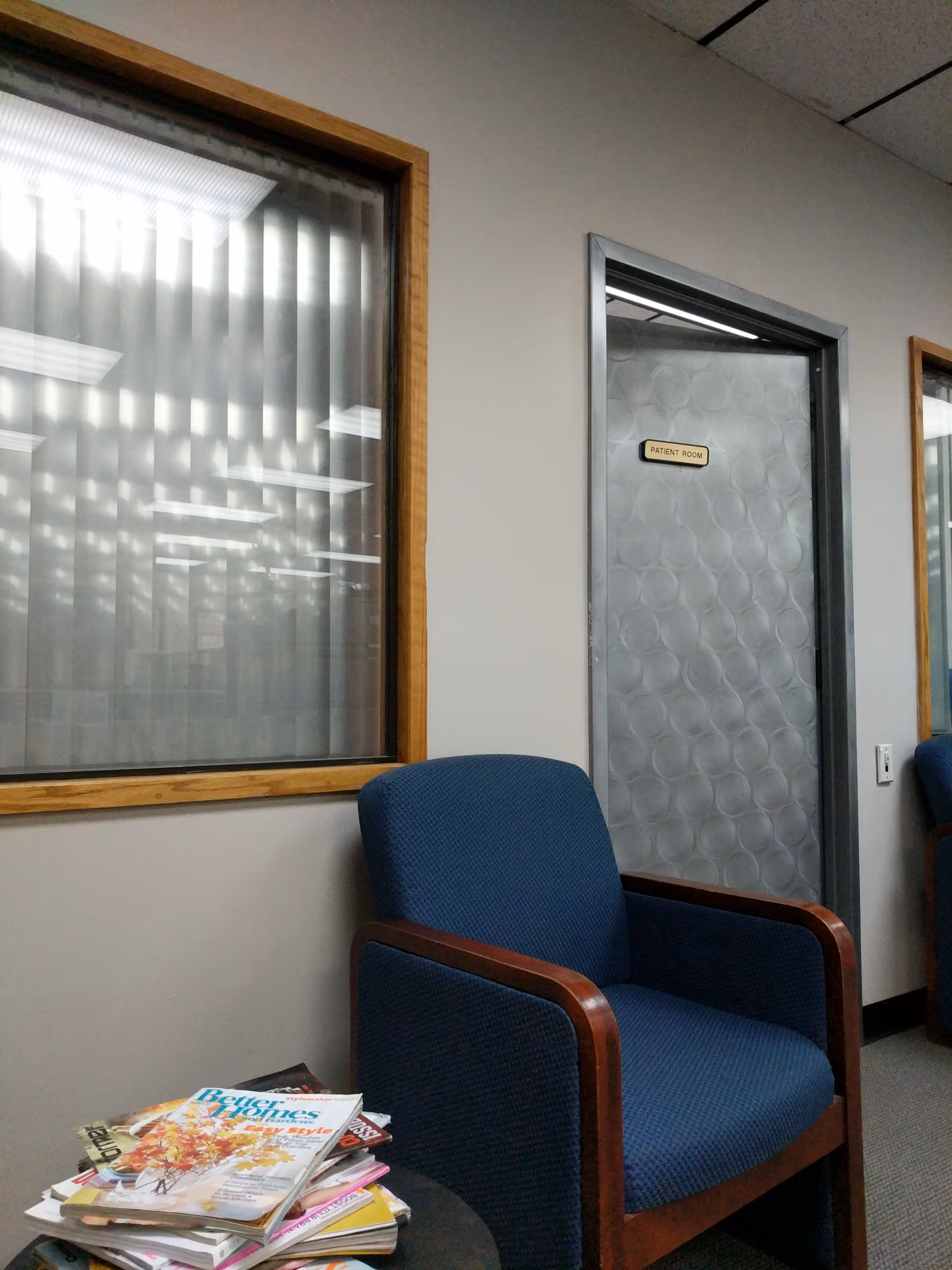 Blue cushioned chair beside a stack of magazines in a hallway near a patient room.
