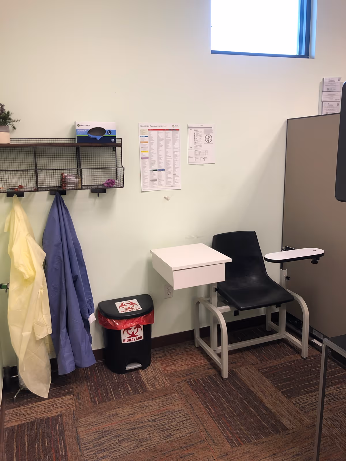 A small medical testing area with a black chair, hanging coats, and posters on the wall.