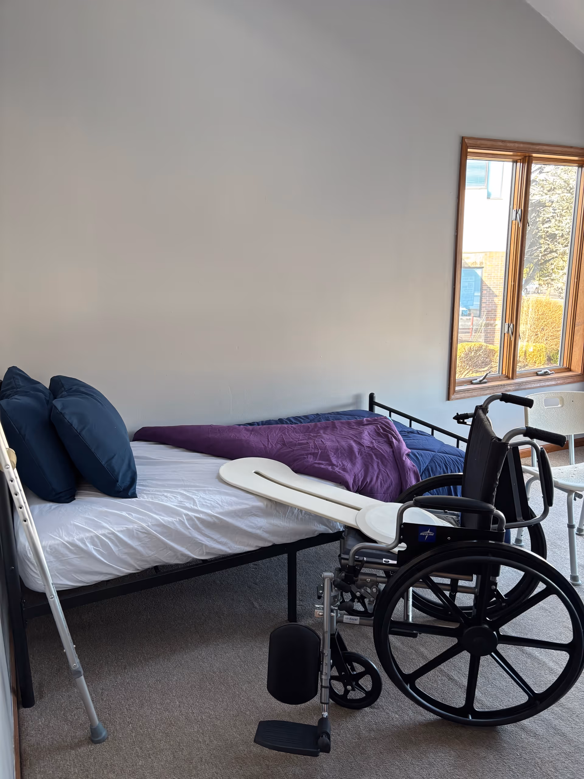 Bedroom with a bed, wheelchair, walker, and window in a senior care facility.
