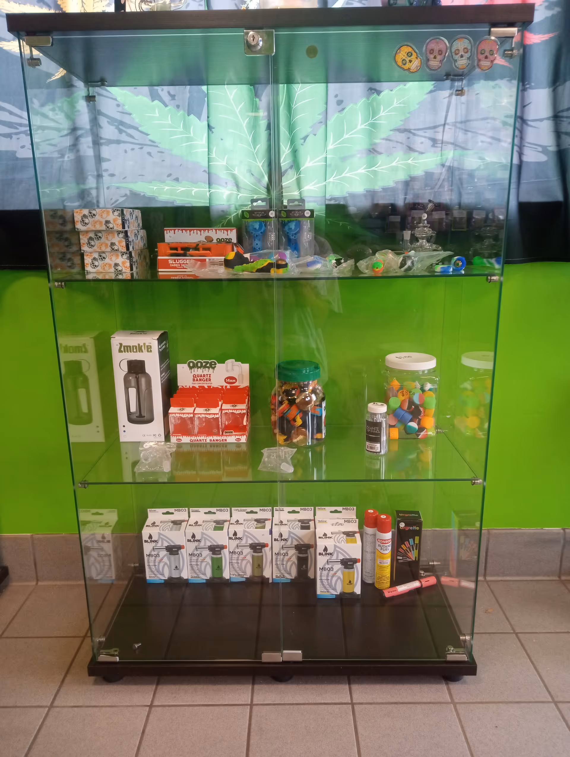 A glass display cabinet filled with assorted products and containers against a bright green wall.