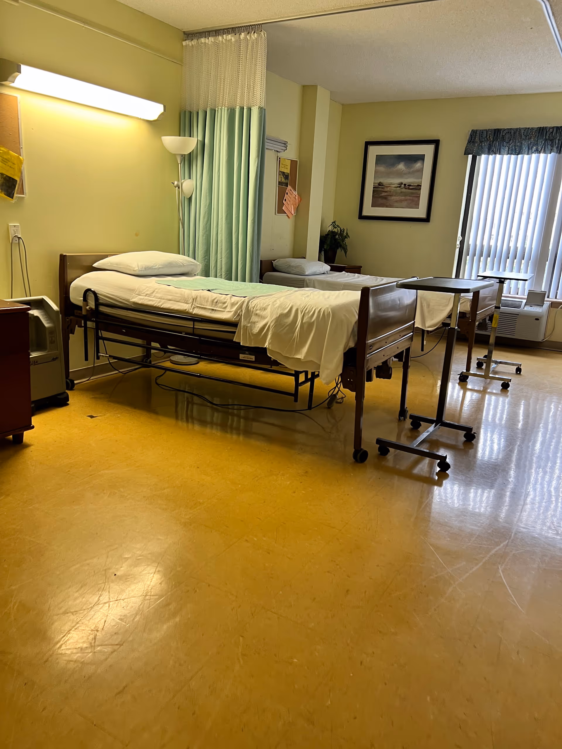 Two hospital beds with green curtains, bedside tables and a window in a patient room.