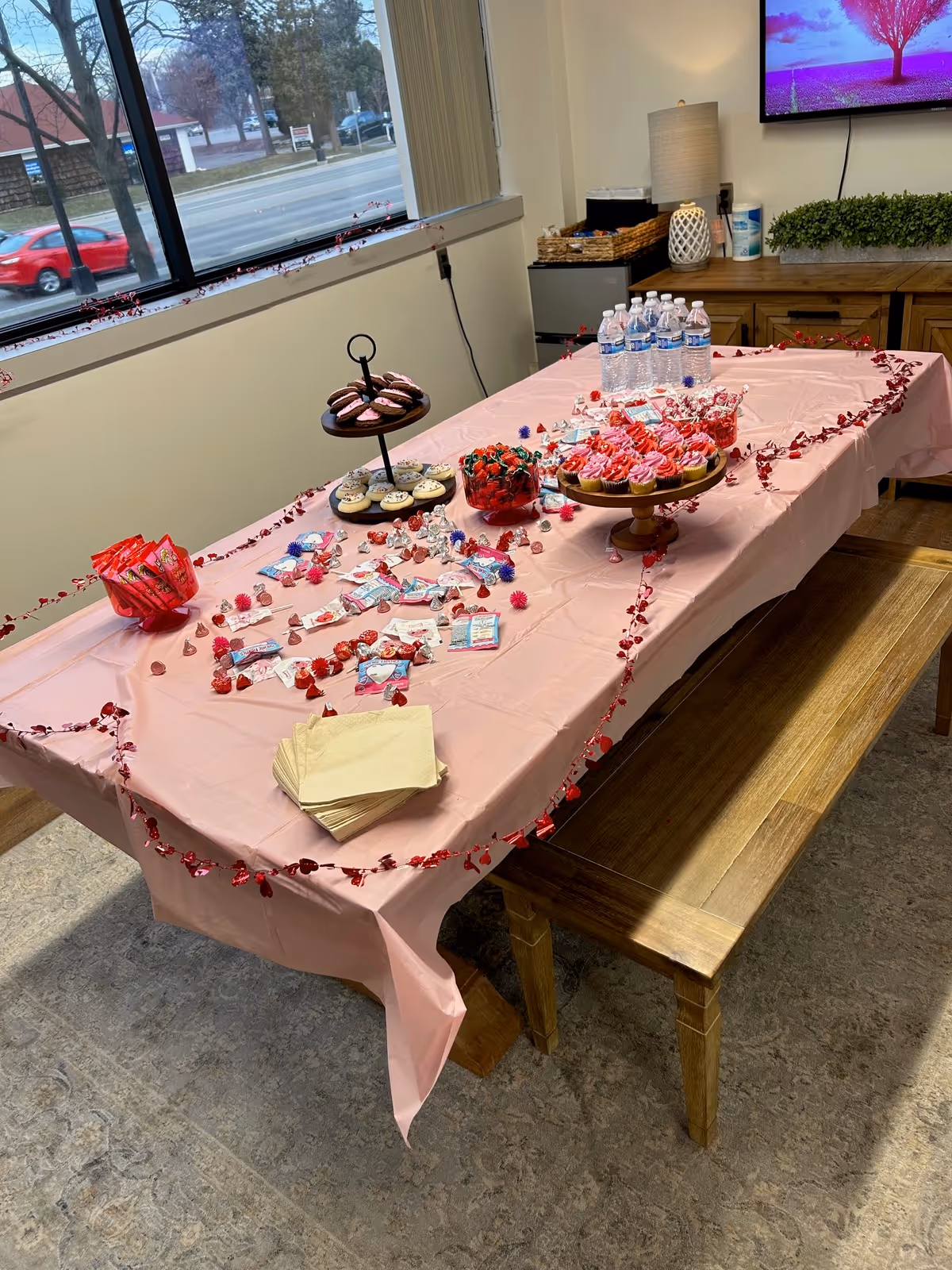 Long dining table with pink tablecloth, cupcakes, candy, and bottled water in a senior living facility.